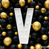 A large white marquee letter shaped like a V is surrounded by a dense arrangement of gold and black balloons for party decor.