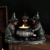A dark metal figurine of three witch cats with pointed hats surrounding a lit candle on a round base.