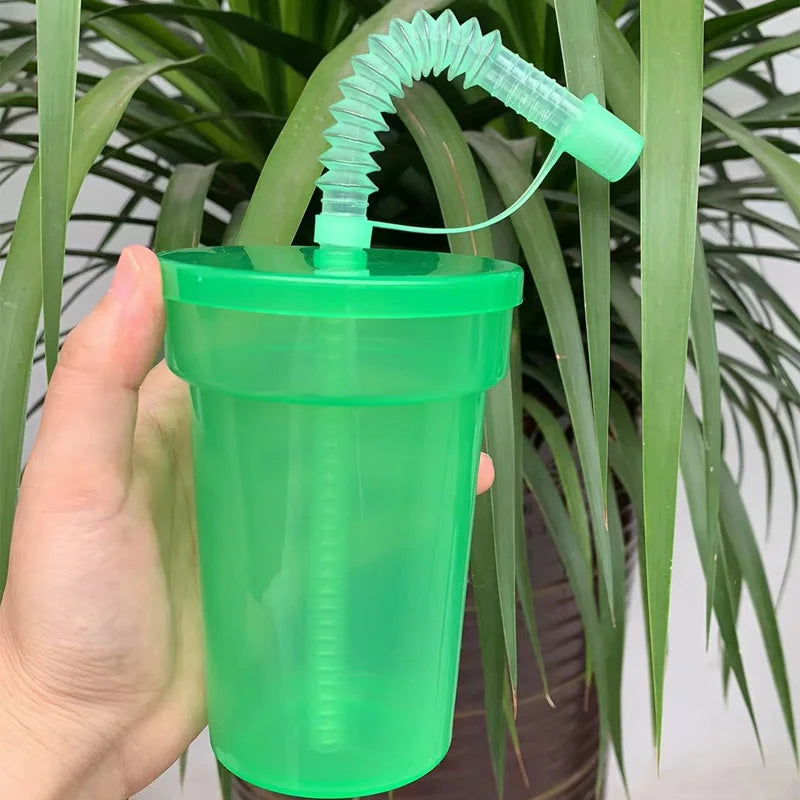 A hand holds a neon green reusable sipper cup with a straw and lid against a background of green foliage.