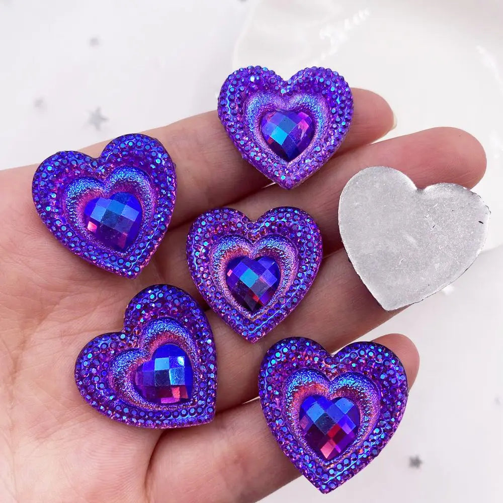 A hand holds a set of twelve heart-shaped iridescent purple and blue peacock resin rhinestone flatbacks.
