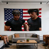 Two men in black shirts stand in front of an American flag backdrop in a gym setting.