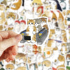 A hand holds a sticker featuring three cats sitting inside a dark gray box.