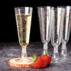 A glass of sparkling champagne next to several clear, elegant disposable champagne glasses and a strawberry on a wooden board