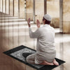 A man in a white thawb and cap is praying on a black prayer mat indoors.