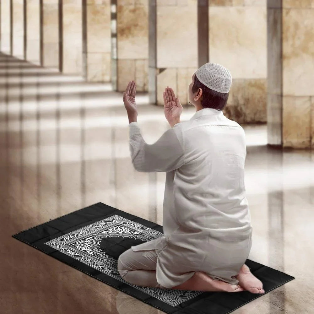 A man in a white thawb and cap is praying on a black prayer mat indoors.