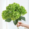 A hand holds a vibrant green artificial hydrangea silk flower bouquet against a white background.
