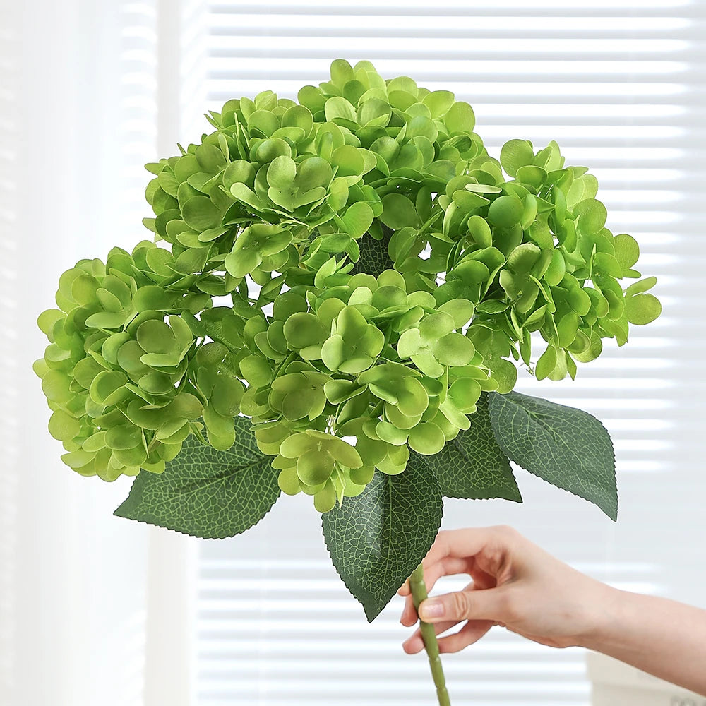 A hand holds a vibrant green artificial hydrangea silk flower bouquet against a white background.