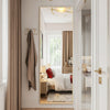 A full length floor mirror with an aluminum frame reflects a bedroom interior with white bedding and light wood furniture.