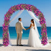 A bride and groom stand under a floral archway made of purple and pink vines on a beach.