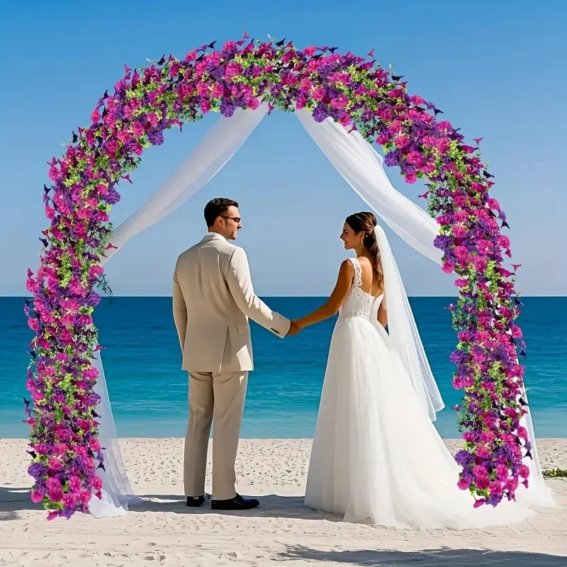 A bride and groom stand under a floral archway made of purple and pink vines on a beach.