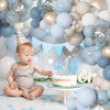 A baby is seated in front of a balloon arch decorated with dusty blue and sand white balloons and confetti accents.