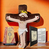 A plush doll depicting Jesus on a cross with a crown and beard, set against a warm orange background next to a Bible.