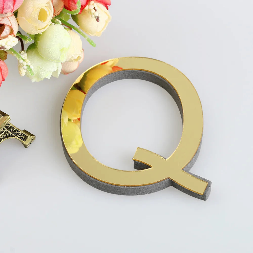 A gold letter Q mirror wall decal set with a textured finish is shown next to a bouquet of pink and cream flowers.