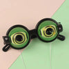 A pair of funny crazy eye costume glasses with black frames and green lenses resting on a light pink and green background.