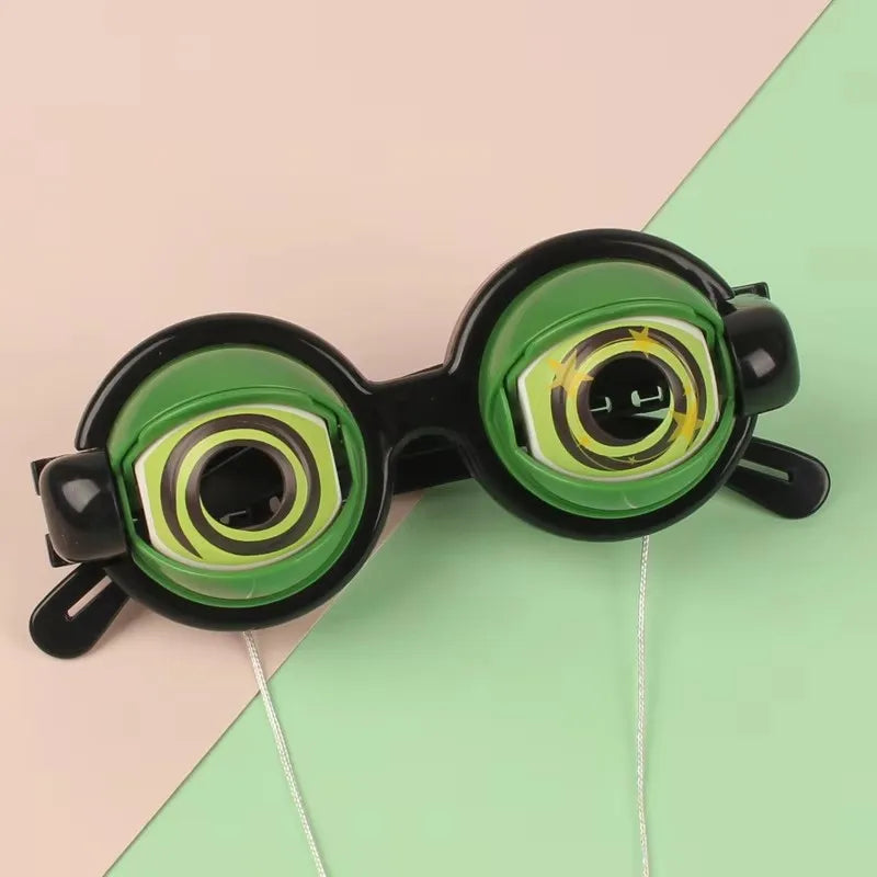 A pair of funny crazy eye costume glasses with black frames and green lenses resting on a light pink and green background.