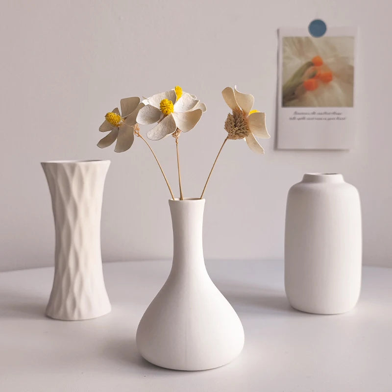 A white ceramic bud vase holds dried flowers on a white surface.