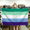 A person holds up a horizontal Pride flag banner with six colored stripes: green, white, light blue, white, blue, and purple.
