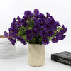 A bouquet of artificial purple pansy flowers with green eucalyptus stems in a light colored pot.