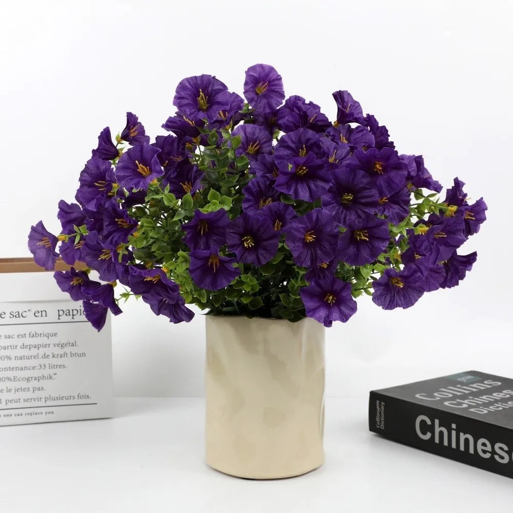 A bouquet of artificial purple pansy flowers with green eucalyptus stems in a light colored pot.