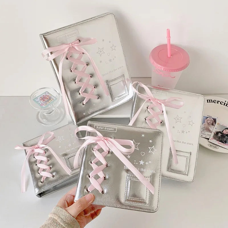 A hand holds a silver leather photo album with pink ribbon ties, surrounded by similar albums and a pink straw cup.