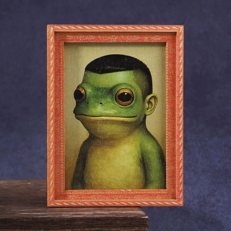 A framed illustration of a green frog face in a miniature photo frame.