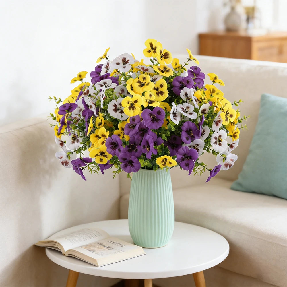 A bouquet of artificial pansy flowers in yellow, purple, and white arranged in a light green vase on a white table.