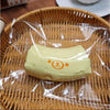 A pale yellow, squishy toy shaped like a banana bear is sealed in clear plastic and placed on a woven bamboo tray.