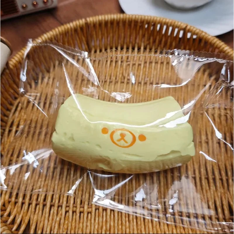 A pale yellow, squishy toy shaped like a banana bear is sealed in clear plastic and placed on a woven bamboo tray.