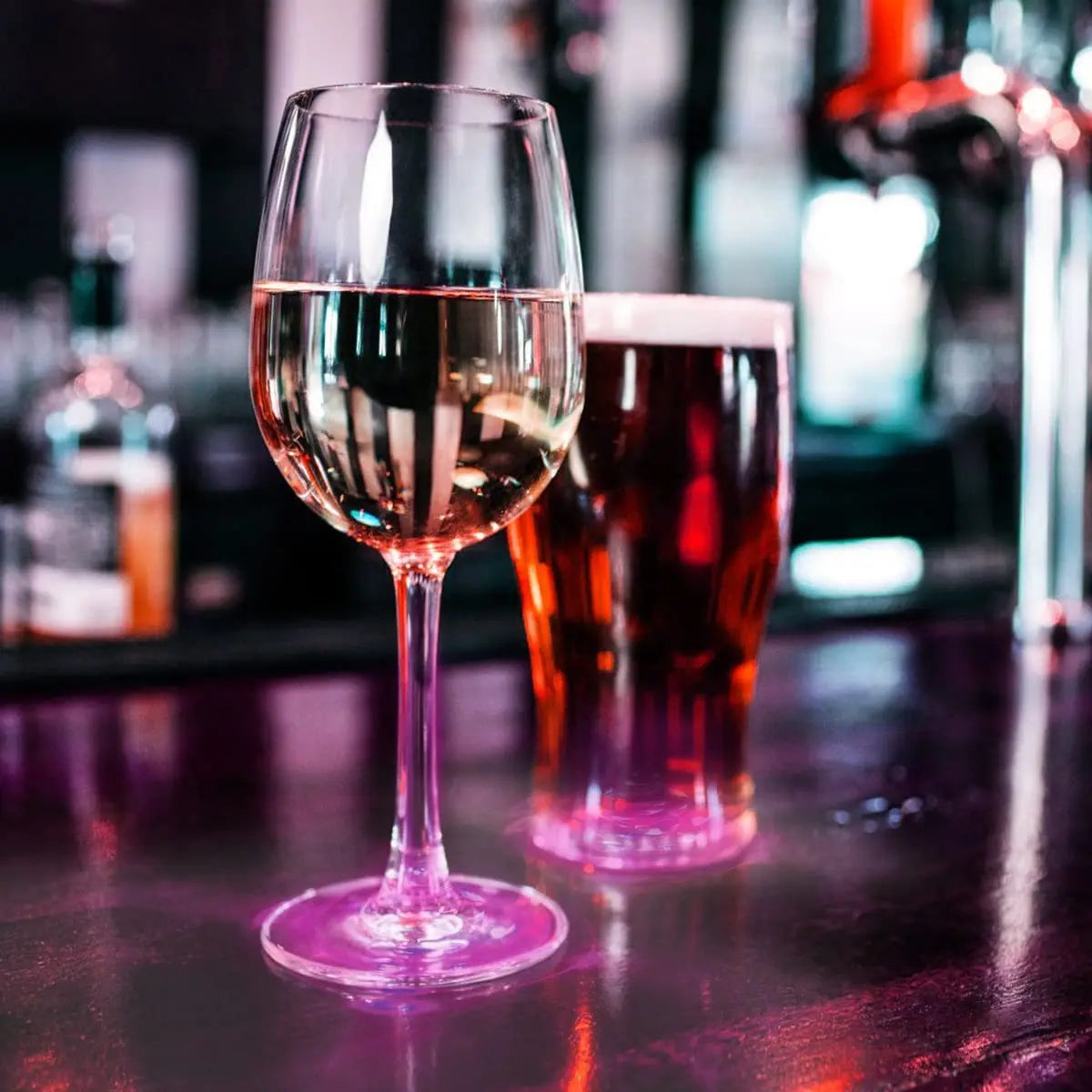 A glass of rosé wine sits next to a glass of beer on a bar counter with soft lighting.