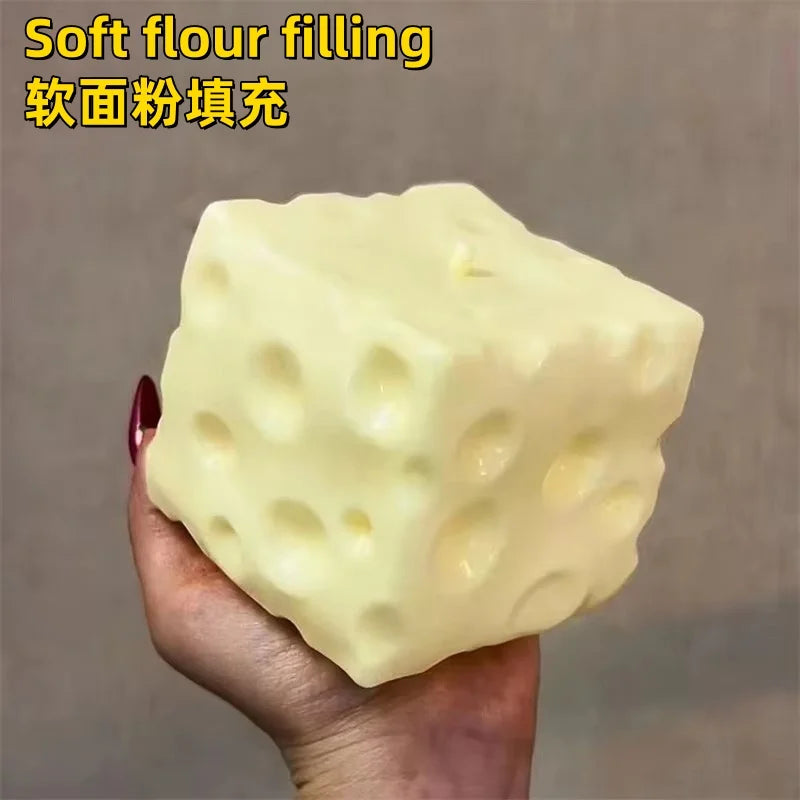 A hand holds a large, pale yellow cheese cube stress ball with visible holes.
