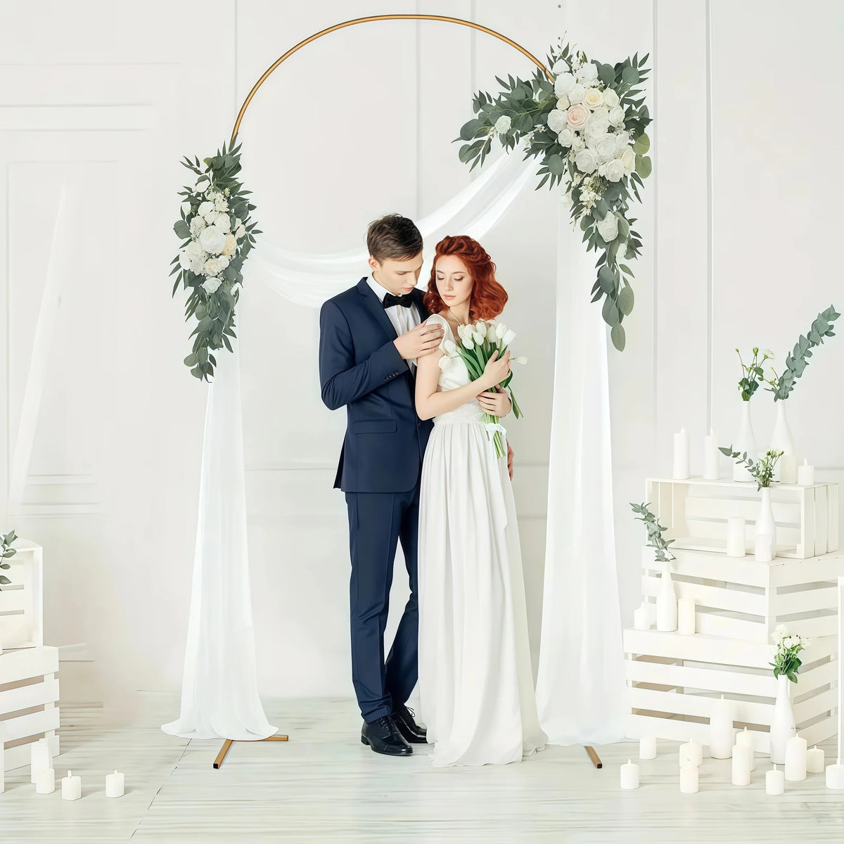 A bride and groom stand beneath an elegant gold metal wedding arch with white floral greenery backdrop.