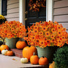 An artificial autumn floral decoration set featuring bright orange and yellow flowers in green pots next to pumpkins.