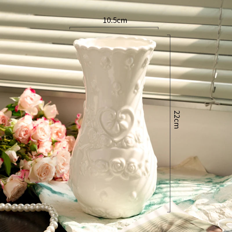 A white ceramic vase with a textured design sits on a striped cloth next to pink roses.