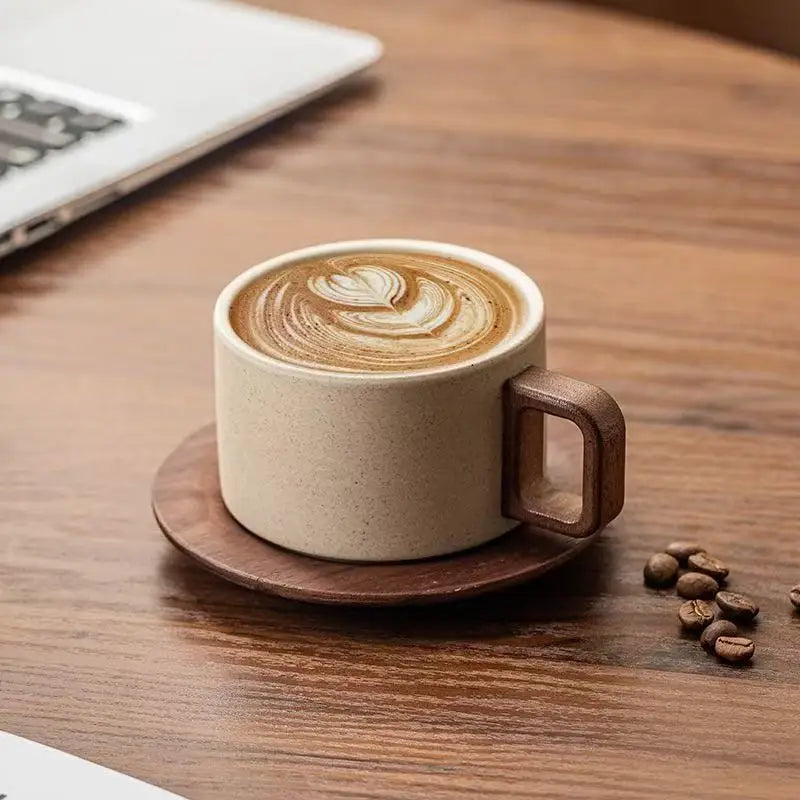 A latte with intricate foam art is served in a beige ceramic coffee cup and saucer on a wooden table.