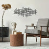 A black geometric metal wall art sculpture hangs on a white wall above a wooden side table and an armchair.