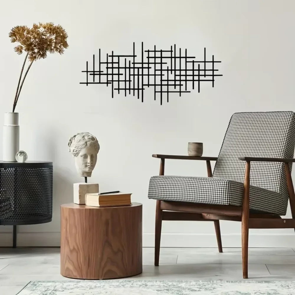 A black geometric metal wall art sculpture hangs on a white wall above a wooden side table and an armchair.