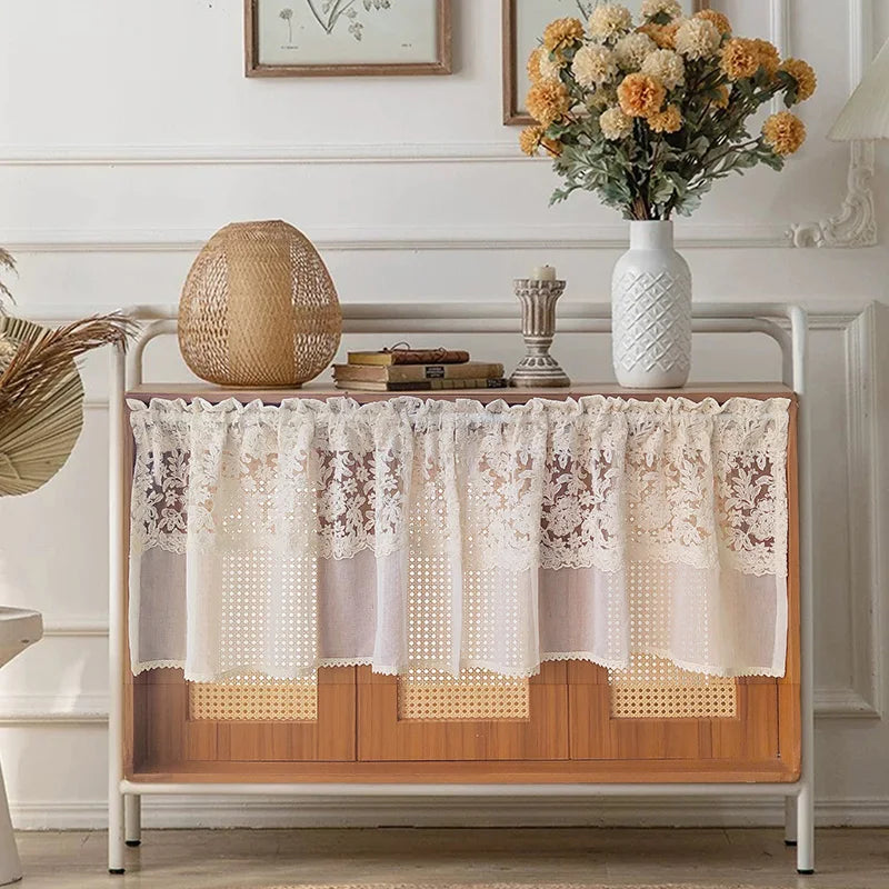 A beige sheer curtain with floral embroidery draped over a wooden shelf in a bright room.