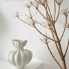 A white minimalist ceramic vase is displayed on a white shelf next to a dry, intricate branch structure.