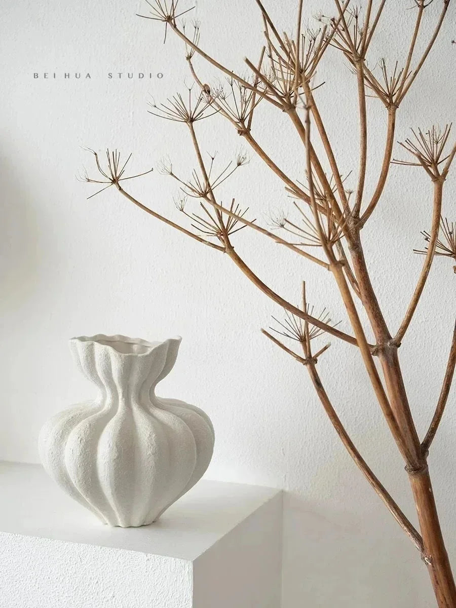 A white minimalist ceramic vase is displayed on a white shelf next to a dry, intricate branch structure.
