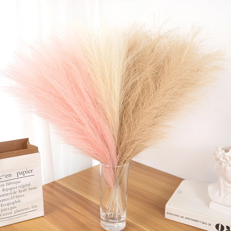 Un ramo de pampas artificial con plumajes rosa y beige en un jarrón de vidrio transparente.