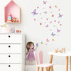 A young girl is interacting with butterfly wall stickers on a white wall next to a pink shelf and white furniture.