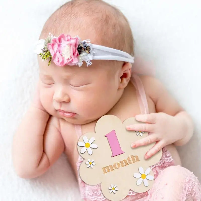 A newborn baby wearing a floral headband, lying down, with wooden monthly milestone cards visible.