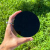 A hand holds a black obsidian mirror reflecting the night sky in a lush green outdoor setting.