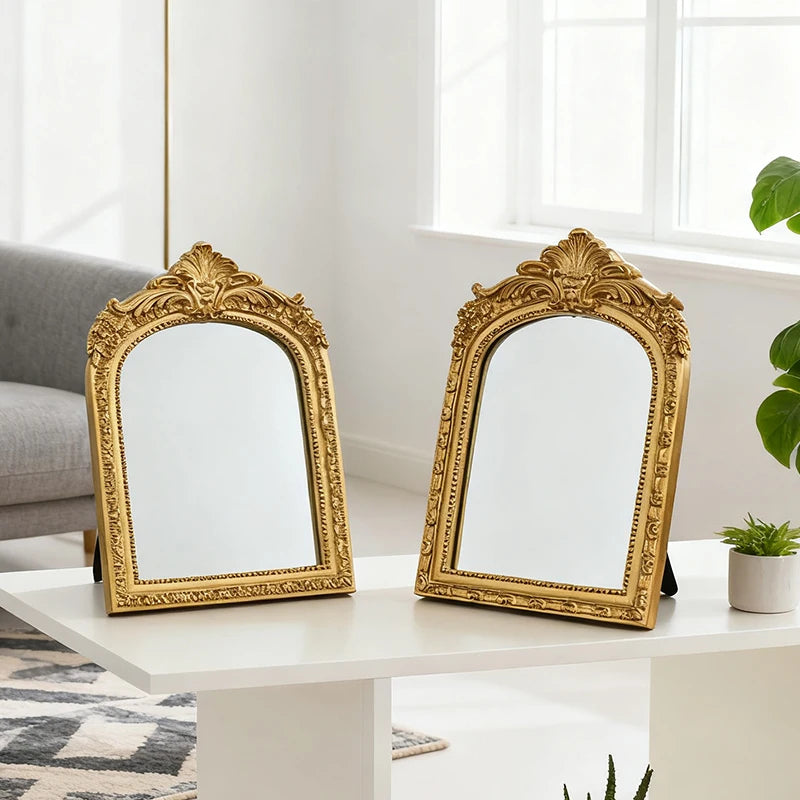 A pair of ornate gold-framed vanity mirrors with arched carvings resting on a white table.