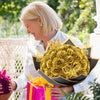 A woman holds a large bouquet of golden artificial roses in an open black box and a pink gift box.