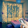 A hand holds a remote control for USB-powered LED curtain lights illuminating sheer curtains with warm glowing lights.