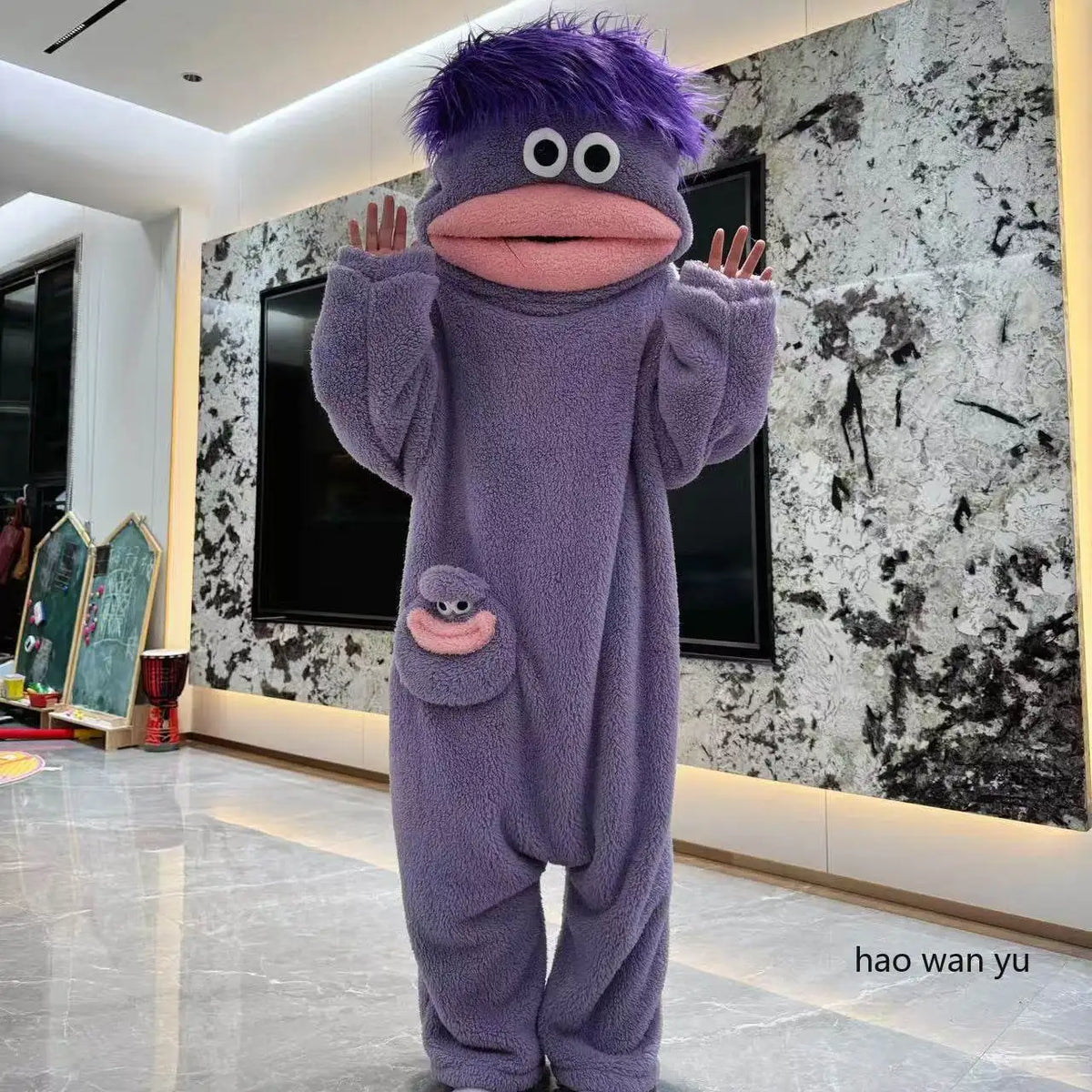 A purple monster coral fleece hooded sleepsuit with large eyes and a pink mouth standing in a modern room.