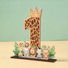 A wooden party ornament shaped like the number one featuring a leopard print and a crown, surrounded by small animal figures.