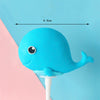 A bright blue whale-shaped cake topper with a white stick, shown against a light blue and pink background.