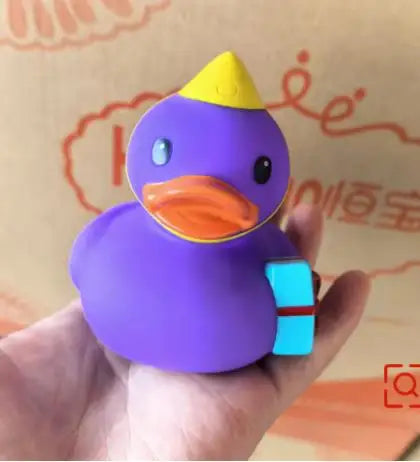 A purple rubber duck bath toy with an orange beak and a yellow crown is held in a hand.